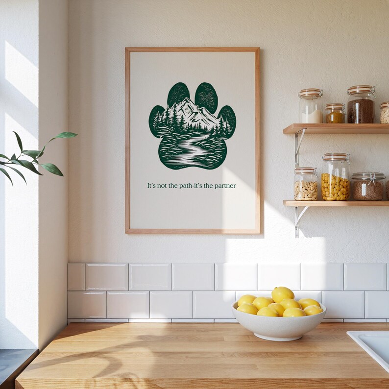 May include: A framed print with a dark green paw print design filled with a mountain and forest landscape. The text "It's not the path-it's the partner" is below the image. The print is displayed on a white wall.