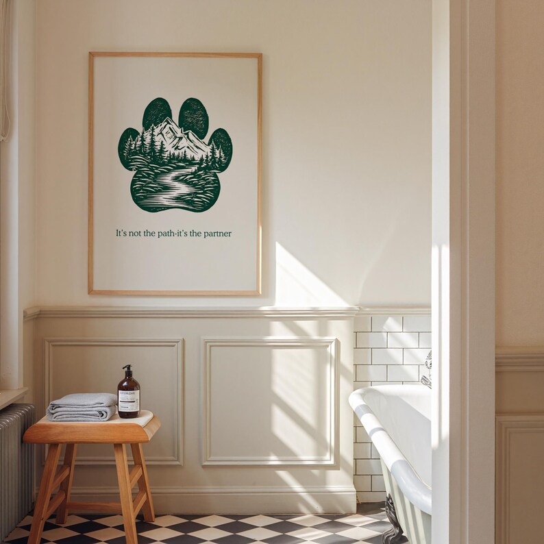 May include: A framed print with a green paw print design, incorporating a mountain and river landscape. The text reads "It's not the path-it's the partner". The artwork is displayed in a bathroom with a wooden stool and a bathtub.