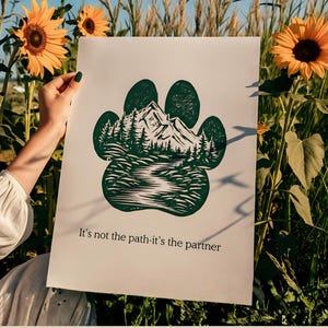 May include: A white poster with a green paw print design, depicting a mountain and forest scene. The text "It's not the path-it's the partner" is printed at the bottom. The poster is held in a field of sunflowers.