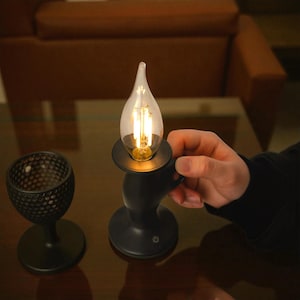 Puede incluir: Lámpara negra con forma de vela con una bombilla transparente en forma de llama. La lámpara tiene una base redonda y un asa. También se ve un segundo objeto decorativo negro con una superficie texturizada.