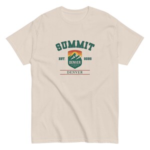 K&ouml;nnte beinhalten: Ein cremefarbenes T-Shirt mit einem Grafikdesign. Das Design beinhaltet das Wort "SUMMIT" in gr&uuml;nem, bogenf&ouml;rmigem Text, eine Berglandschaft und den Text "DENVER" und "8088". Das T-Shirt hat eine klassische Passform.
