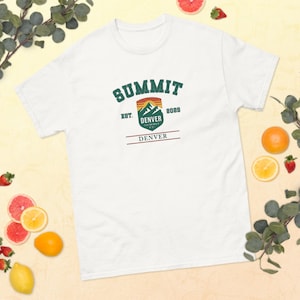 May include: White t-shirt with a green, orange, and brown graphic that reads "SUMMIT DENVER" with the text "EST. 2022" and a mountain design. The shirt is made of a soft material and is suitable for casual wear.