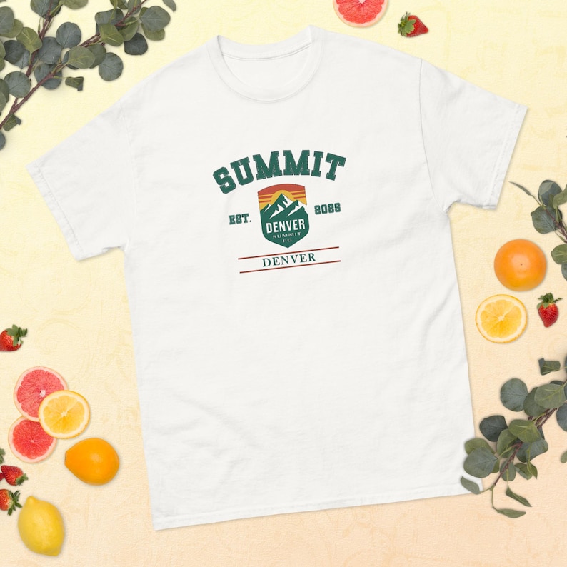 K&ouml;nnte beinhalten: Wei&szlig;es T-Shirt mit einem gr&uuml;nen, orangefarbenen und braunen Aufdruck mit der Aufschrift "SUMMIT DENVER", dem Text "EST. 2022" und einem Bergdesign. Das Shirt besteht aus einem weichen Material und eignet sich f&uuml;r Freizeitkleidung.