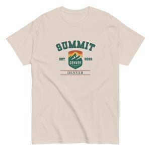 K&ouml;nnte beinhalten: Cremefarbenes T-Shirt mit einem Grafikdesign. Das Design enth&auml;lt das Wort "SUMMIT" in gr&uuml;nem, bogenf&ouml;rmigem Text, eine Berglandschaft und den Text "DENVER" und "8088". Das T-Shirt hat einen Rundhalsausschnitt und kurze &Auml;rmel.