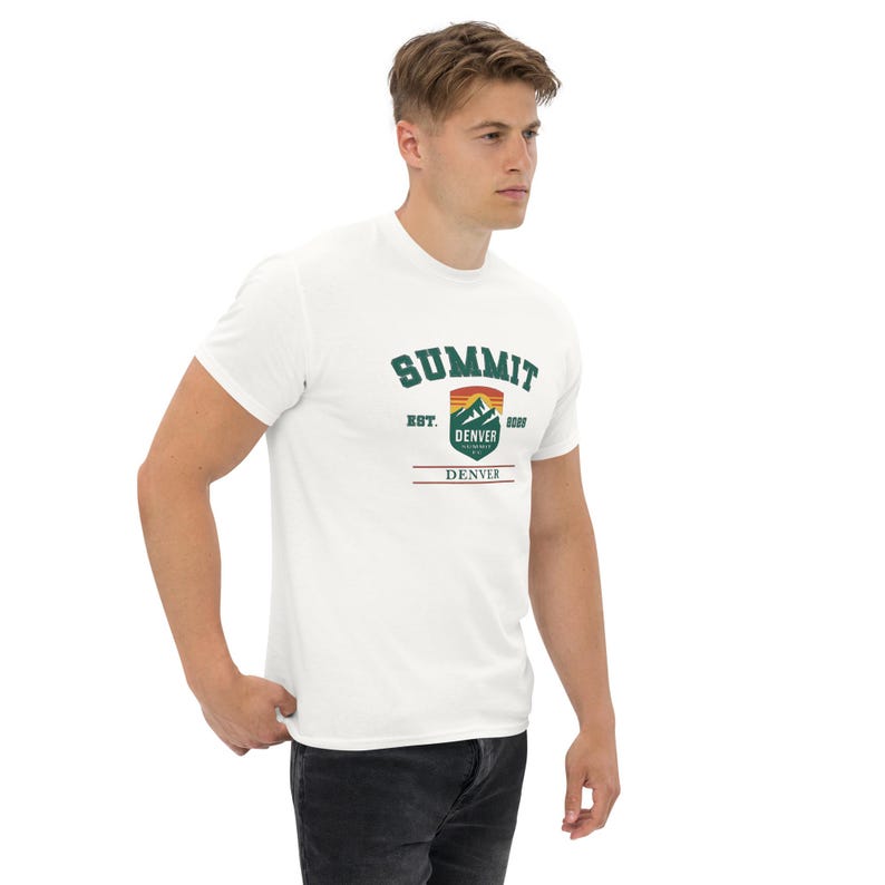 K&ouml;nnte beinhalten: Wei&szlig;es T-Shirt mit einem gr&uuml;nen, orangefarbenen und braunen Aufdruck mit der Aufschrift "SUMMIT DENVER" und dem Jahr 1889. Das T-Shirt hat kurze &Auml;rmel und einen Rundhalsausschnitt. Die Person auf dem Bild tr&auml;gt eine dunkelblaue Jeans.
