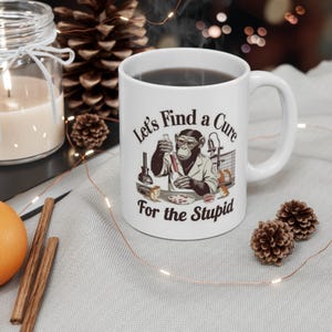 May include: White ceramic mug with a black liquid inside. The mug features a graphic of a chimpanzee in a lab coat with the text "Let's Find a Cure For the Stupid".