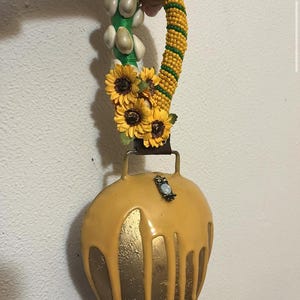 May include: A decorative bell with a yellow and gold finish, adorned with sunflowers and an owl charm. The handle is decorated with yellow and green beads, shells, and a green fabric. The bell is held by a hand.