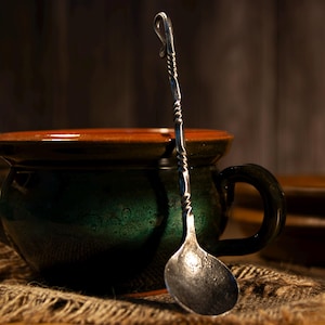 Puede incluir: Una taza de cerámica verde oscuro con borde marrón y una cuchara decorativa de plata. La cuchara, con mango retorcido, se apoya en la taza, evocando una bebida caliente y reconfortante.