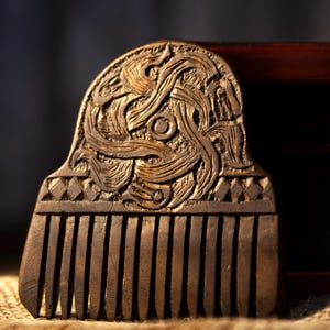May include: A wooden comb with intricate carvings. The comb features a detailed design on the handle, with vertical teeth extending downwards. The wood has a warm, brown tone, and the comb rests on a textured surface.