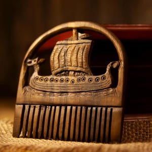 May include: A wooden comb with a carved Viking ship design. The comb is brown and features a detailed depiction of a longship with a sail, set against an arched frame. The comb rests on a burlap surface.