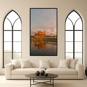 May include: A framed print depicting the Golden Temple in Amritsar, India, reflected in water. The image has a warm colour palette, with the temple illuminated against a sunset sky. The print is displayed above a white sofa.