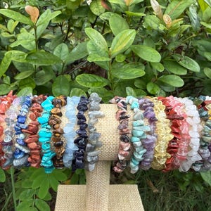 May include: A display of colorful gemstone chip bracelets. The bracelets are arranged on a tan fabric-covered stand, showcasing a variety of colors including red, blue, green, and yellow. The background features lush green foliage.