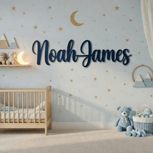 Könnte beinhalten: Ein Kinderzimmer in Hellblau und Goldtönen. An der Wand befindet sich ein marineblaues Namensschild mit der Aufschrift "Noah James". Das Zimmer umfasst ein Holzbett, Teddybären und dekorative Elemente. Die Wände sind mit Monden und Sternen verziert, was eine beruhigende Umgebung schafft.