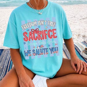 May include: A light blue t-shirt with the text "FREEDOM ISN'T FREE SACRIFICE WE SALUTE YOU" in red, white, and blue. The shirt has a patriotic theme, with stars and stripes design elements. The shirt is worn on a beach.