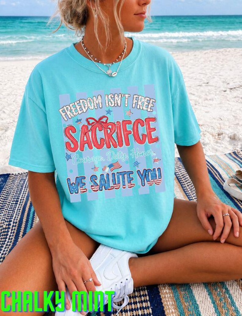 May include: A light blue t-shirt with the text "FREEDOM ISN'T FREE SACRIFICE WE SALUTE YOU" in red, white, and blue. The shirt has a patriotic theme, with stars and stripes design elements. The shirt is worn on a beach.
