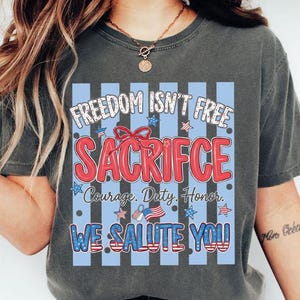 May include: Dark gray t-shirt with a patriotic design. The graphic includes the text "FREEDOM ISN'T FREE SACRIFICE" in red and white, with blue stripes and stars. Additional text reads "Courage. Duty. Honor. WE SALUTE YOU!"