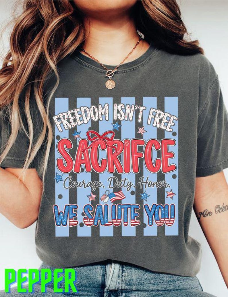 May include: Dark gray t-shirt with a patriotic design. The graphic includes the text "FREEDOM ISN'T FREE SACRIFICE" in red and white, with blue stripes and stars. Additional text reads "Courage. Duty. Honor. WE SALUTE YOU!"