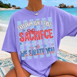 May include: A violet-colored t-shirt with the text "FREEDOM ISN'T FREE SACRIFICE" in red, white, and blue. The shirt also features the words "Courage, Duty, Honor" and "WE SALUTE YOU". The word "VIOLET" is printed in green at the bottom.