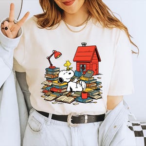Puede incluir: Camiseta color crema con un diseño de Snoopy relajándose sobre libros con Woodstock, una caseta roja y una lámpara roja. También incluye una taza roja. La camiseta es una prenda casual para el día a día.