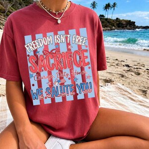 May include: A maroon t-shirt with the text "FREEDOM ISN'T FREE SACRIFICE" in a patriotic design. The shirt also includes the phrases "Courage, Duty, Honor" and "WE SALUTE YOU!!" The shirt is worn on a beach.