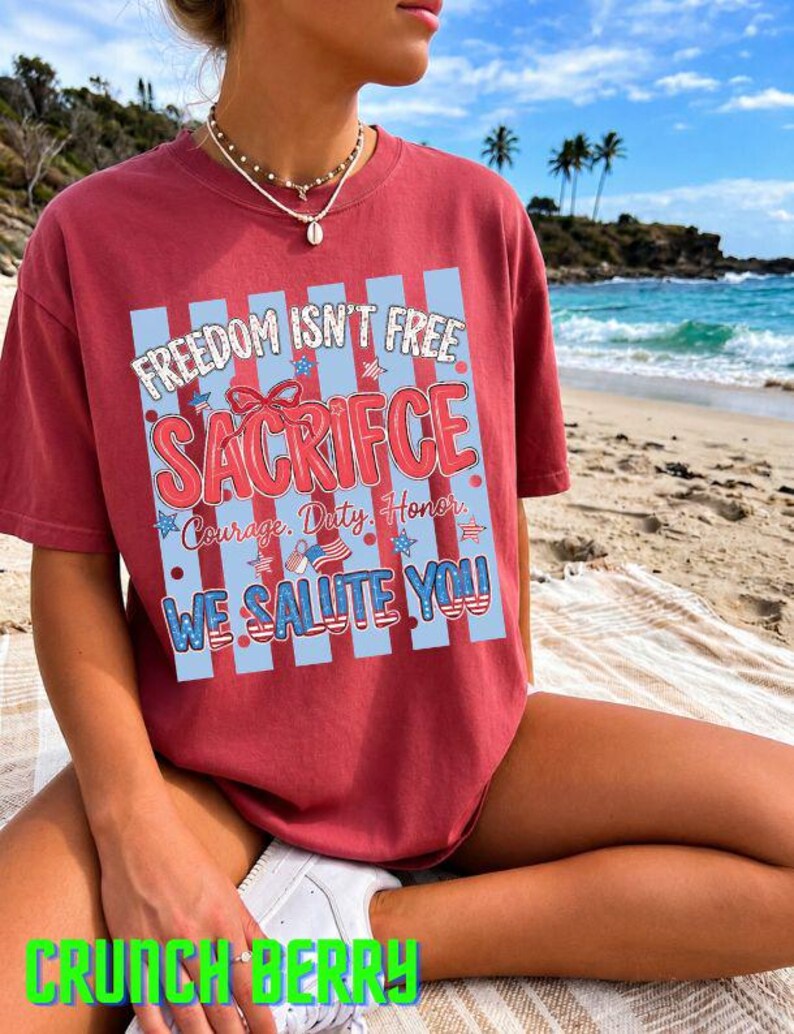 May include: A maroon t-shirt with the text "FREEDOM ISN'T FREE SACRIFICE" in a patriotic design. The shirt also includes the phrases "Courage, Duty, Honor" and "WE SALUTE YOU!!" The shirt is worn on a beach.