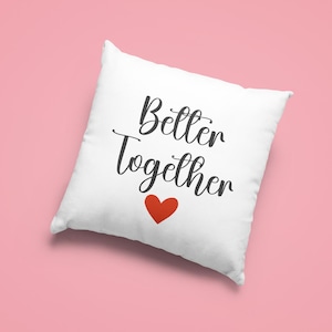 May include: White square pillow with the words "Better Together" in black script and a red heart. The pillow is on a pink background. A decorative item for a bedroom or living room.