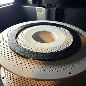 May include: Close-up of a white perforated circular object with a black textured ring. The object is part of a 3D printer, with the printer head visible in the background. The object has a series of small holes.