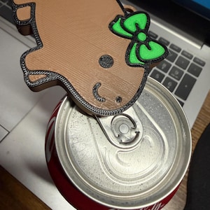 以下が含まれることがあります： 緑のリボンが付いたジンジャーブレッドマンの形をした、茶色の3Dプリントのドリンク缶トッパー。赤いコカ・コーラ缶の上に置かれています。缶はシルバーで、コカ・コーラのロゴが入っています。