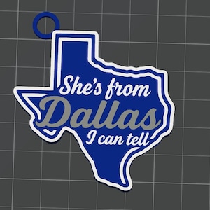 May include: A blue and white Texas-shaped keychain with a circular ring at the top. The text "She's from" and "I can tell" is printed in white, while the word "Dallas" is printed in grey across the center of the state-shaped design.