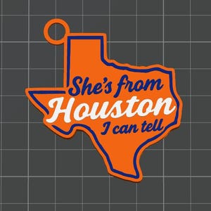 May include: An orange keychain shaped like the state of Texas with a blue border and a circular loop at the top. The text "She's from Houston I can tell" is printed across the center in a combination of blue script and white block lettering.