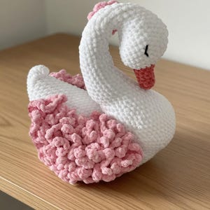 Könnte beinhalten: Ein handgefertigter, weiß-rosa gehäkelter Schwan. Der Schwan hat einen gebogenen Hals und einen rosa Schnabel. Der Körper ist weiß mit rosa Akzenten. Der Schwan sitzt auf einer Holzoberfläche.