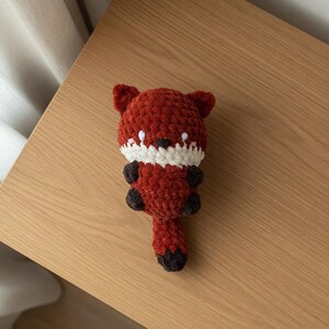 May include: A handmade crochet fox toy in shades of red, white, and brown. The fox has a rounded body, small ears, and a fluffy tail. The toy is lying on a wooden surface.