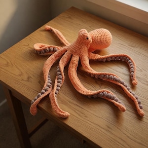Könnte beinhalten: Ein gehäkeltes Oktopus-Spielzeug in Pfirsich- und Grautönen. Der Oktopus hat einen abgerundeten Kopf und acht lange, flexible Tentakel. Die Tentakel sind pfirsichfarben mit grauen Details. Das Spielzeug liegt auf einer Holzoberfläche.