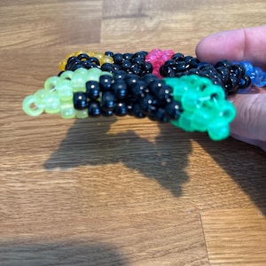 Può includere: Un braccialetto di perline multicolore, con perline nere, verdi, gialle, rosa e blu. Le piccole perline rotonde sono infilate insieme per formare una fascia flessibile. Il braccialetto è mostrato su una superficie di legno.