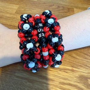 May include: A stack of handmade beaded bracelets in red, black, and white. The bracelets are made of small, round beads strung together. The letters "MCR" are spelled out in black beads. The bracelets are on a wooden surface.
