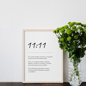 May include: A framed print with the handwritten text "11:11" in black. Below the numbers is a paragraph of text in Spanish. A vase of green flowers sits to the right of the frame on a wooden surface.