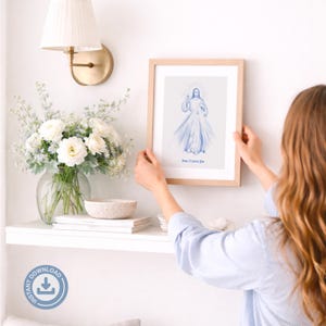 May include: A framed print with a light wood frame. The print features a blue illustration of a religious figure with the text "Jesus, Trust in You". The print is being held up against a white wall.