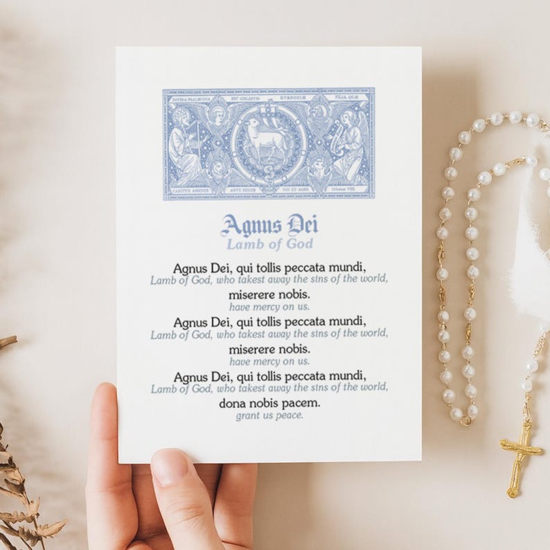 May include: A white card with the text "Agnus Dei, Lamb of God" in blue script. The card features a detailed blue illustration at the top and printed text below. A rosary with a gold cross is visible on the right.