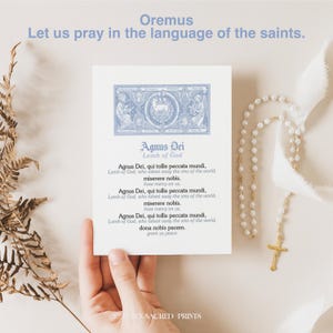 May include: A white card featuring the text "Agnus Dei" and "Lamb of God" in blue script. A hand holds the card, with a rosary and gold cross visible. The words "Oremus" and "Let us pray in the language of the saints" are at the top of the image.