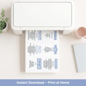 May include: A white printer printing invitation cards with blue decorative borders and text. The cards are being printed on white paper. A small potted plant, a pink mug, and a light blue notebook are on the white surface.
