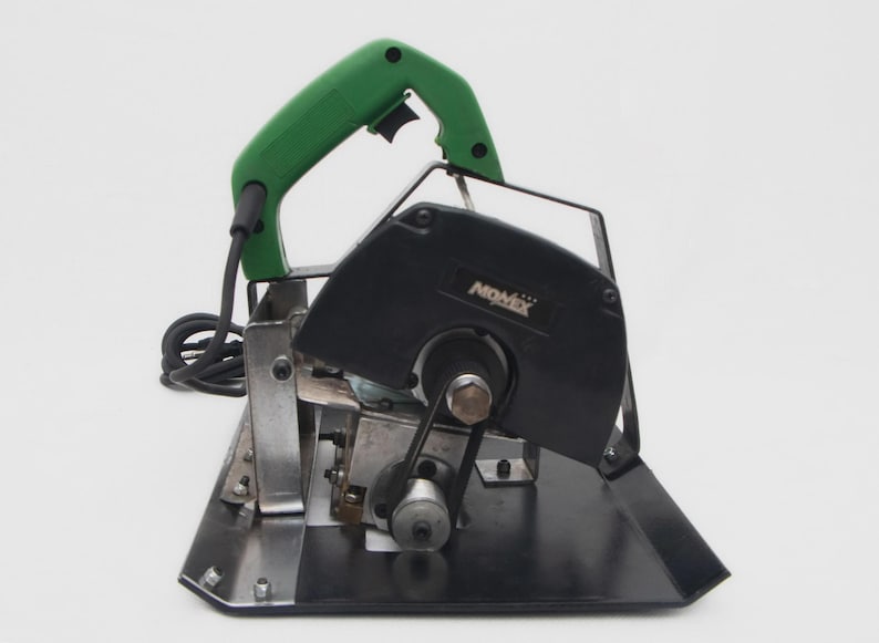 May include: A black and silver Nomex brand tile cutter with a green handle. The power tool has a black blade guard and a silver base. The cord is black.
