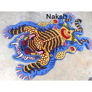 May include: A vibrant, hand-tufted rug in a unique shape, featuring a tiger design. The rug showcases a blue background with yellow, brown, and gray details, and the word "Naksh" is visible. The rug is a decorative floor covering.