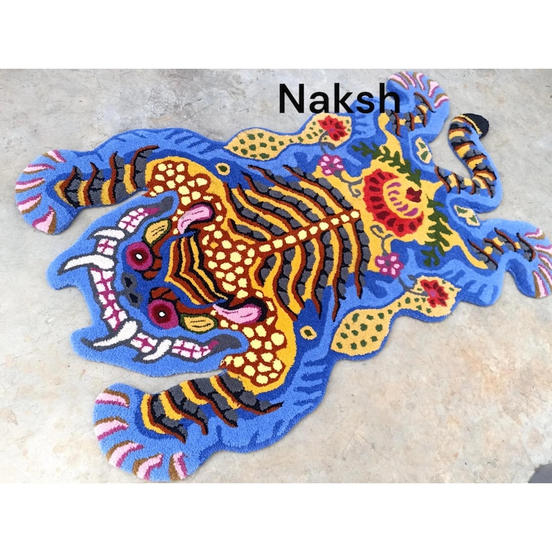 May include: A vibrant, hand-tufted rug in a unique shape, featuring a tiger design. The rug showcases a blue background with yellow, brown, and gray details, and the word "Naksh" is visible. The rug is a decorative floor covering.