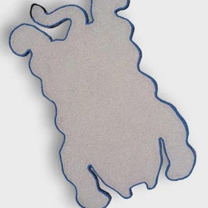 May include: A gray, abstract-shaped rug with a blue border. The rug has a unique, organic form, with a textured surface. The blue border outlines the shape, adding a contrasting detail to the neutral color palette. The rug is designed for home decor.