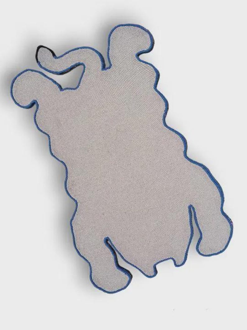 May include: A gray, abstract-shaped rug with a blue border. The rug has a unique, organic form, with a textured surface. The blue border outlines the shape, adding a contrasting detail to the neutral color palette. The rug is designed for home decor.