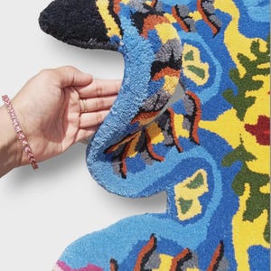 May include: A vibrant, abstract rug in a unique shape, featuring a mix of blue, yellow, orange, and green hues. The rug has a textured, tufted surface and is held by a hand, showcasing its intricate design and color palette. The rug's design is reminiscent of a bird.