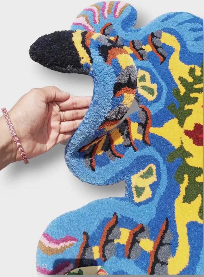 May include: A vibrant, abstract rug in a unique shape, featuring a mix of blue, yellow, orange, and green hues. The rug has a textured, tufted surface and is held by a hand, showcasing its intricate design and color palette. The rug's design is reminiscent of a bird.