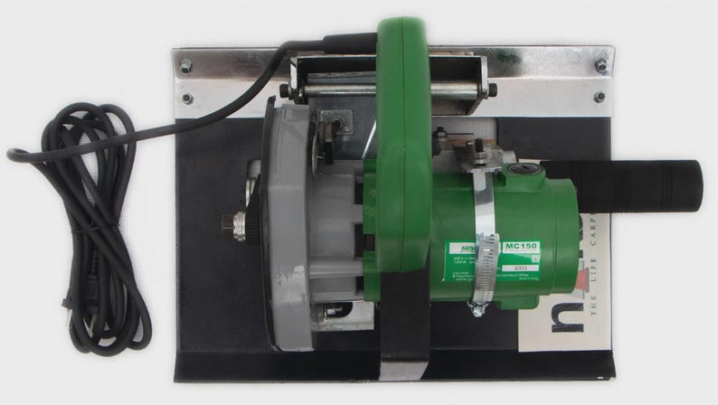 May include: A green and gray power tool with a black handle and cord. The tool is mounted on a black base with a silver metal frame. The tool has a label that reads "MC150".