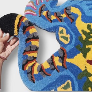 May include: A colorful, abstract rug with a unique shape. The rug features a vibrant mix of blue, yellow, black, orange, and green. The design appears to be hand-tufted, with a soft, textured surface. The rug's irregular shape adds to its artistic appeal.