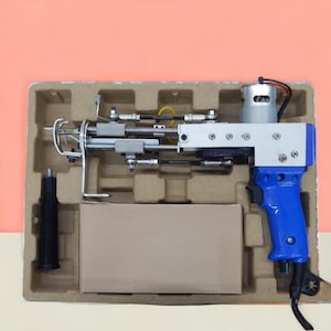 May include: A tufting gun, a tool for creating rugs and other textile art, is shown in its packaging. The gun features a blue handle, a silver body, and various mechanical components. It is presented in a cardboard tray against a peach background.